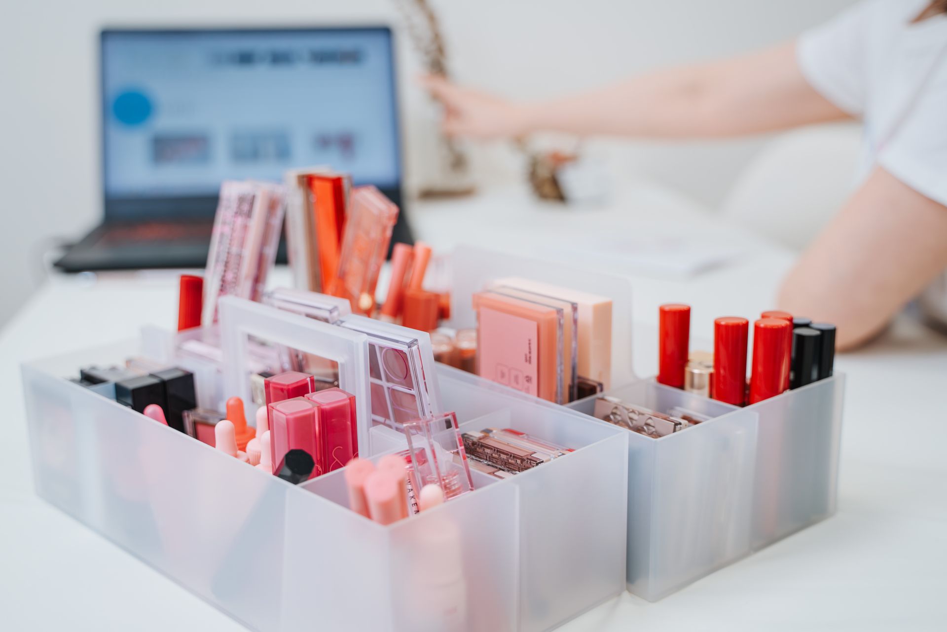 一位女士坐在辦公桌前，手裡拿著一台筆記型電腦和一盤化妝品。