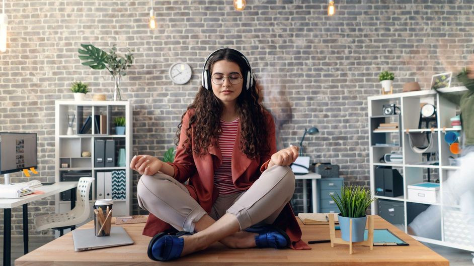 A woman is sitting on a desk in a lotus position with headphones on.