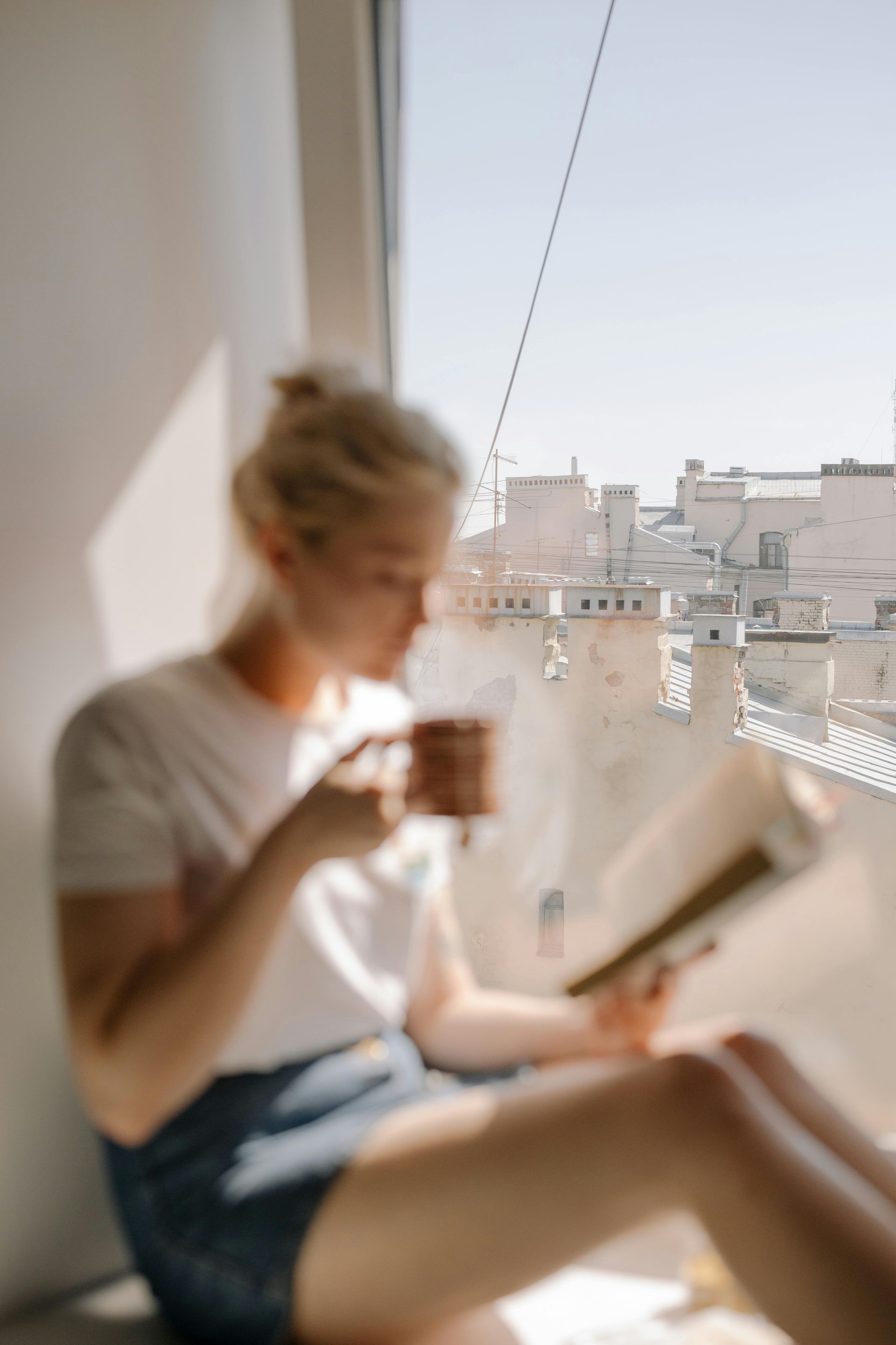 A woman is sitting on a window sill drinking coffee and reading a book.