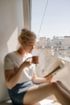 A woman is sitting on a window sill drinking coffee and reading a book.