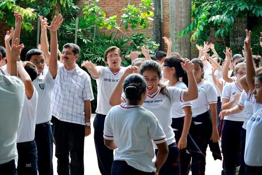 Estudiantes y profesores uniformados, de pie al aire libre con los brazos en alto, participando en una actividad grupal.