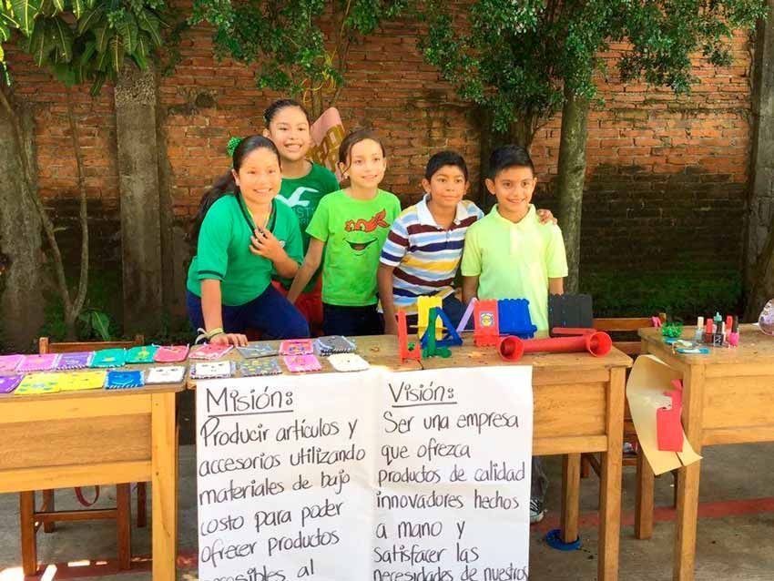 Cinco estudiantes se encuentran detrás de una mesa de aula cubierta de objetos hechos a mano, carteles con la misión y la visión de su proyecto.