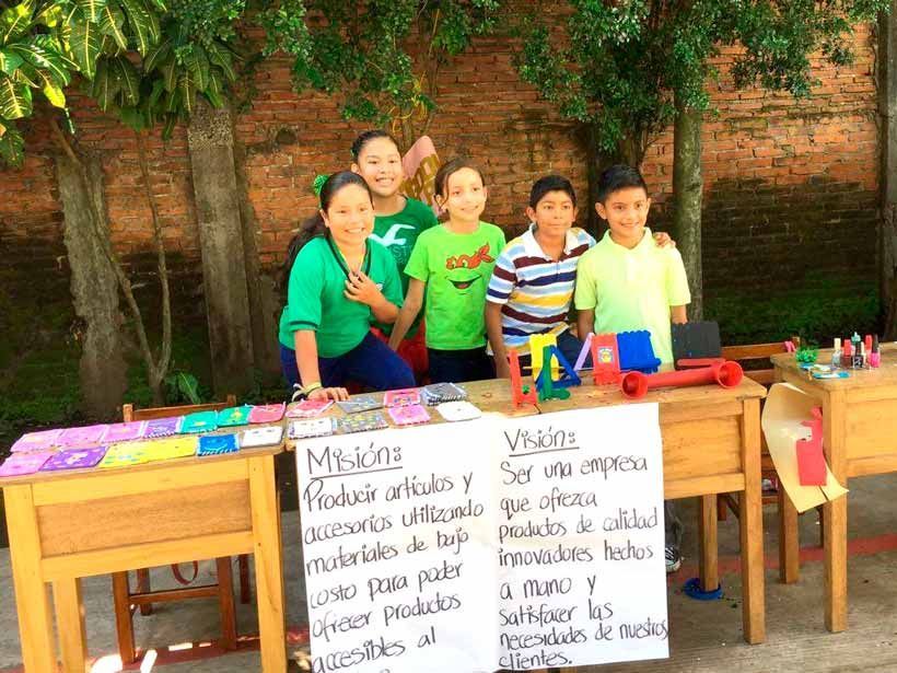 Cinco niños están de pie detrás de una mesa donde se exhiben artículos hechos a mano, con declaraciones de misión y visión escritas en un cartel blanco.