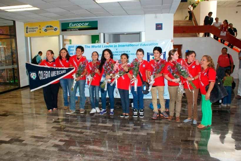 Un grupo de personas vestidas con camisetas rojas se encuentra en un pasillo interior, sosteniendo flores y un banderín frente a una pancarta publicitaria.