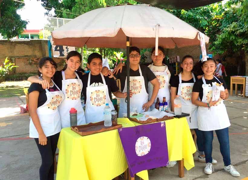 Seis personas con delantales blancos sobre camisas negras están de pie detrás de un puesto de comida cubierto de amarillo, bajo una gran sombrilla beige.