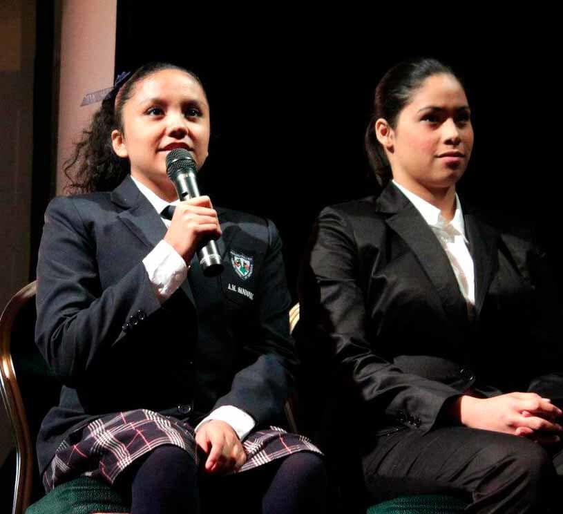 Dos personas con chaquetas formales están sentadas en el escenario. Una de ellas sostiene un micrófono mientras habla; la otra se sienta a su lado.