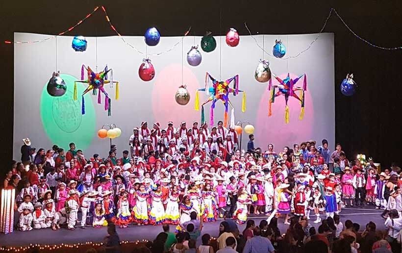 Un numeroso grupo de artistas en el escenario, ataviados con trajes tradicionales y coloridos, bajo piñatas colgantes y adornos decorativos.