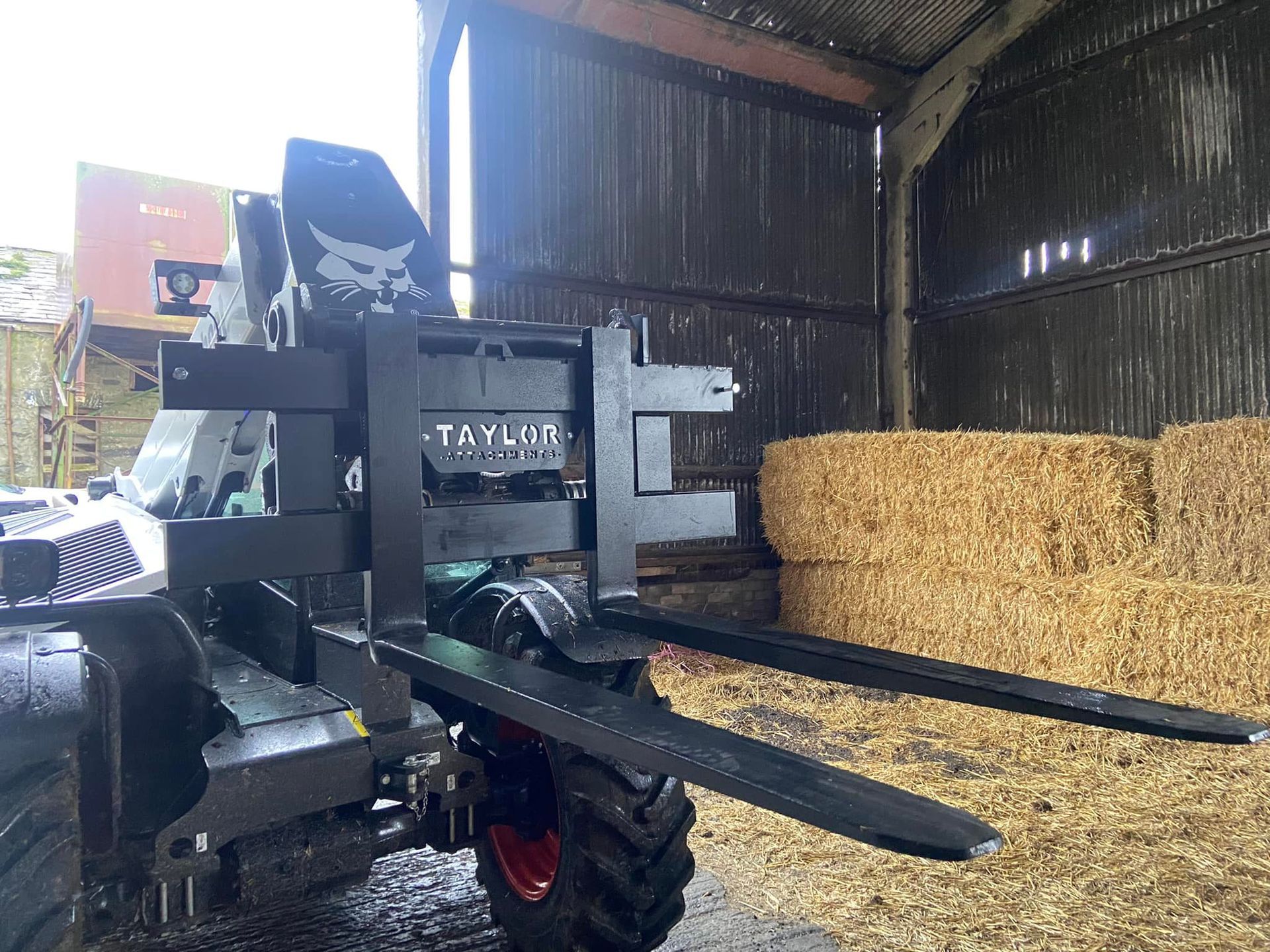 A tractor with a forklift attached to it is in a barn filled with hay bales.