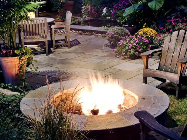 A circular stone fire pit with a bright flame, surrounded by patio furniture and lush garden plants at night.