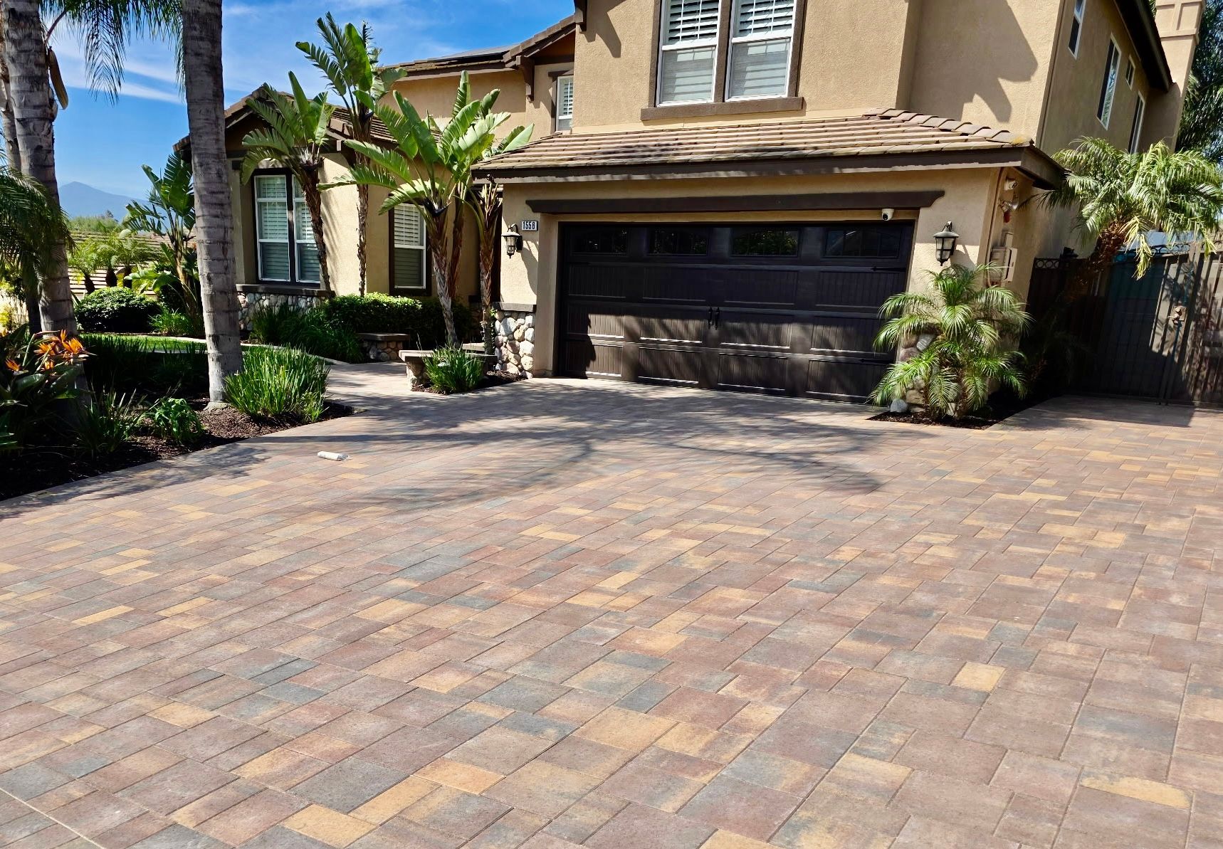 A two-story tan house with a dark garage door and a textured, multi-colored stone paver driveway in a sunny yard.