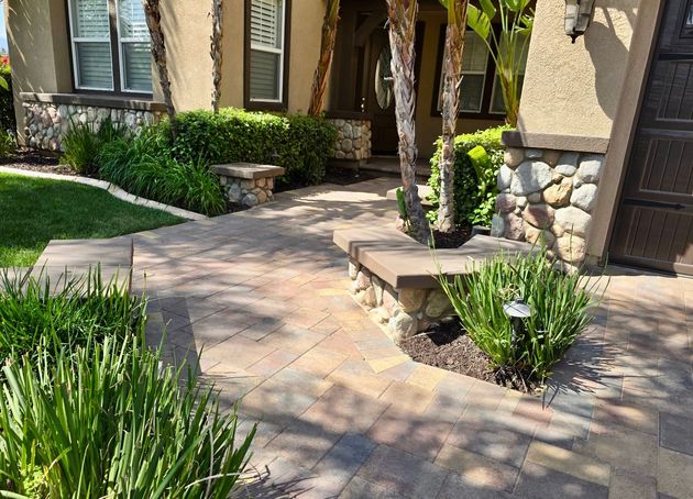A paved walkway leads to a house entrance, flanked by low stone walls, green shrubbery, and two palm trees.