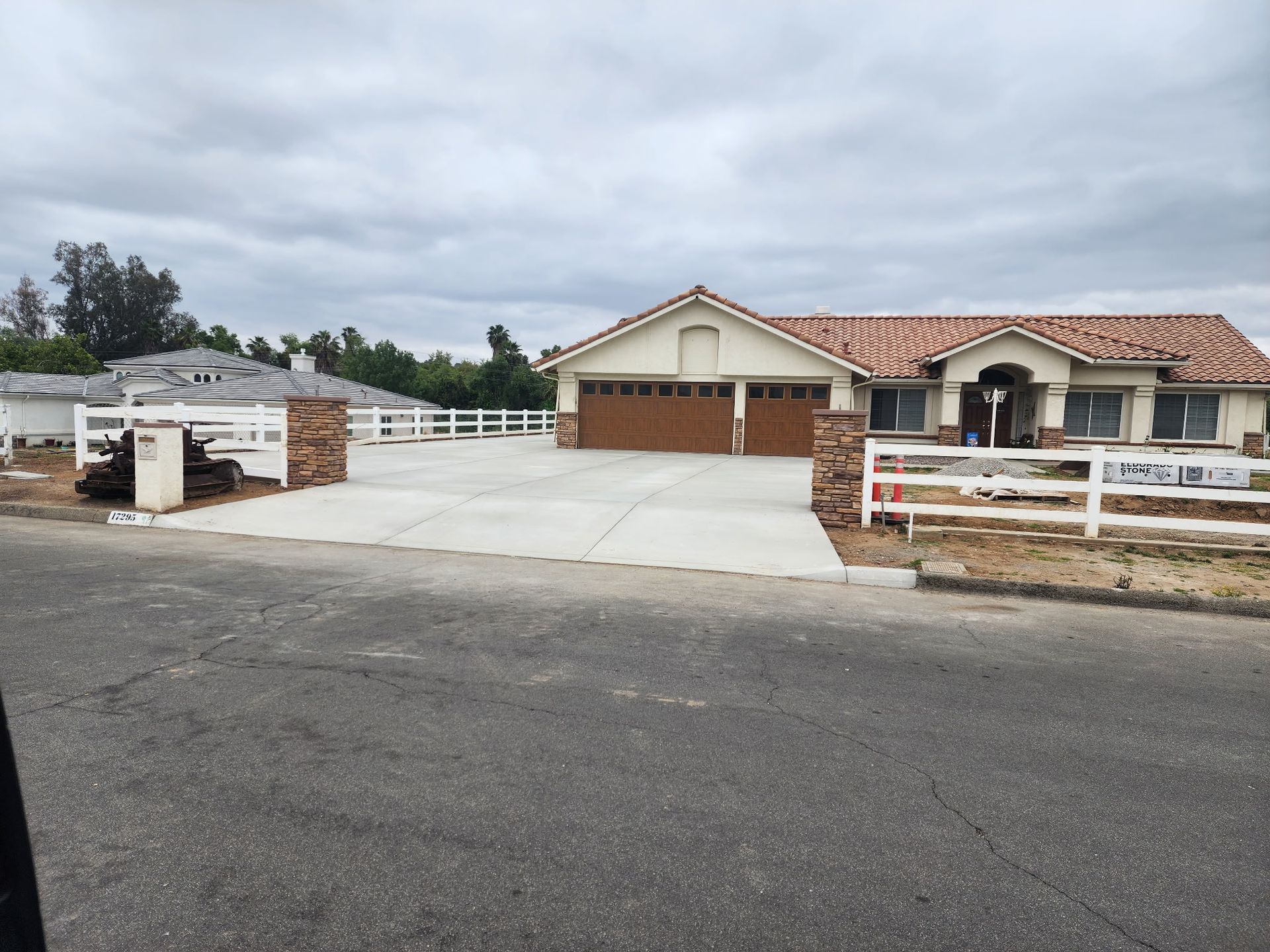A one-story tan house with a red tile roof, three garage doors, and a white fence, seen from an asphalt street.