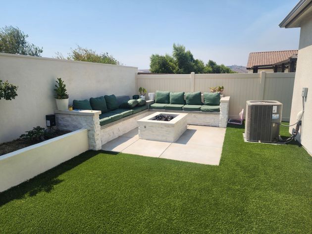An outdoor patio featuring built-in L-shaped benches with green cushions, a stone fire pit, and artificial grass.