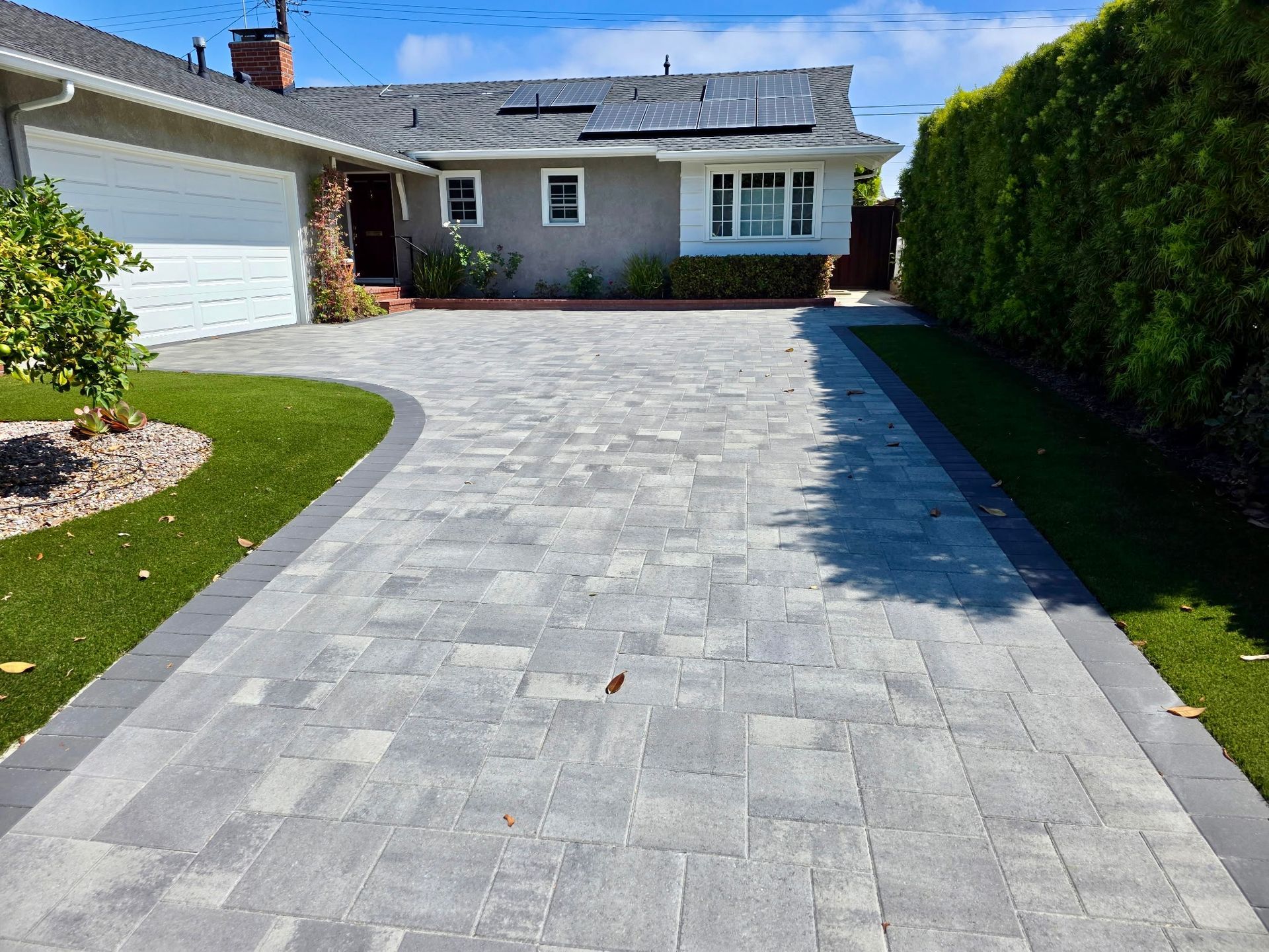 A residential driveway paved with gray stone tiles, bordered by lush green grass and a tall hedge leading to a house.