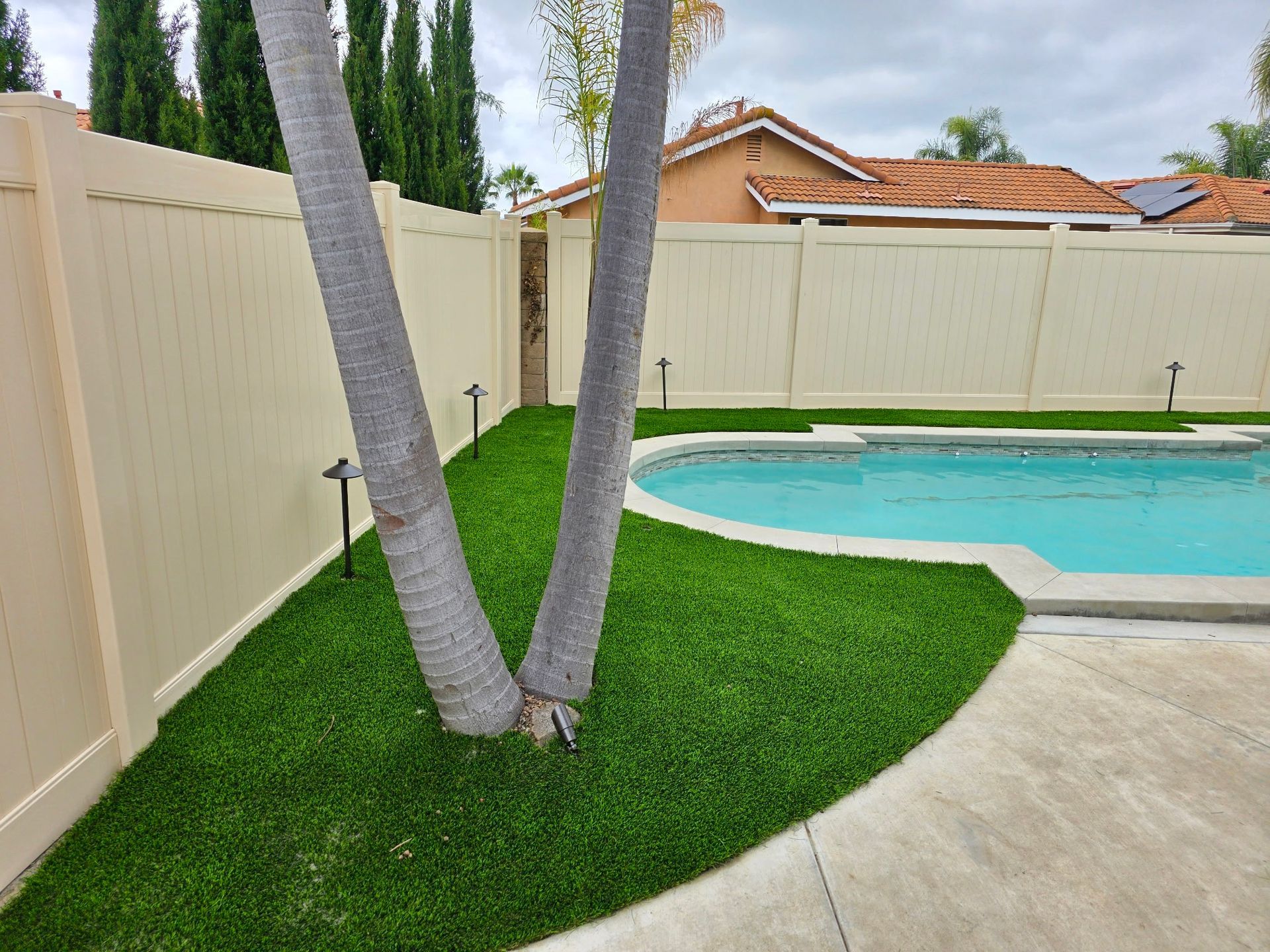 A backyard pool with artificial turf, a V-shaped tree trunk, and a tan vinyl fence under a cloudy sky.