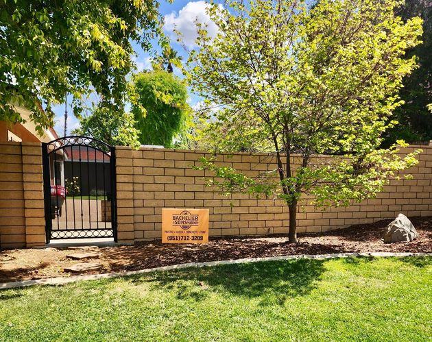 A tan brick wall with a black metal gate, a small tree, and a promotional sign in a landscaped yard.