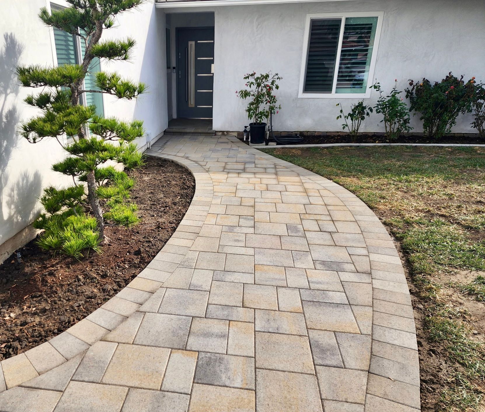 A paved walkway made of rectangular stones leads to the front door of a white house with a manicured shrub and lawn.