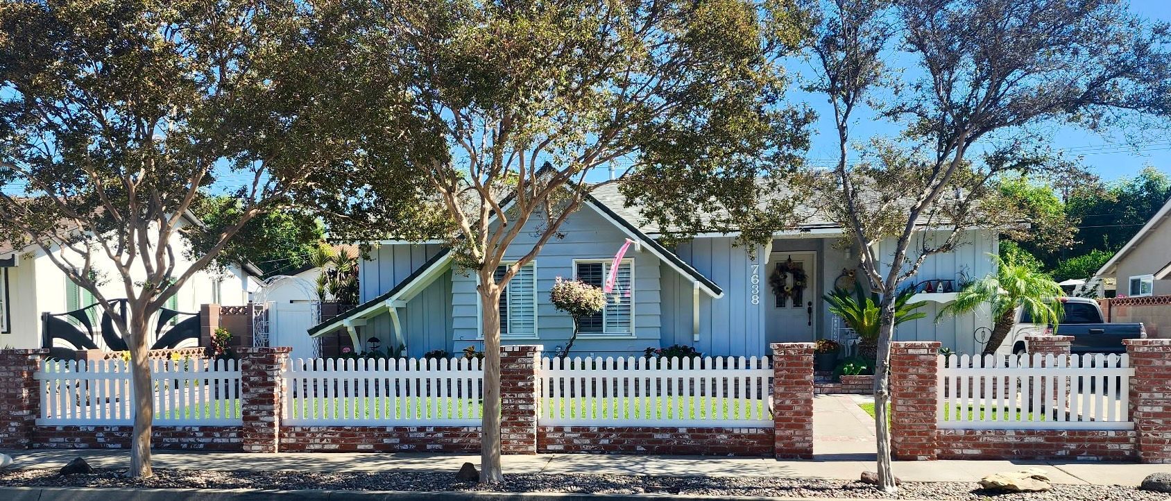A light blue, single-story house behind a white picket fence with brick pillars, shaded by large trees under a clear sky.