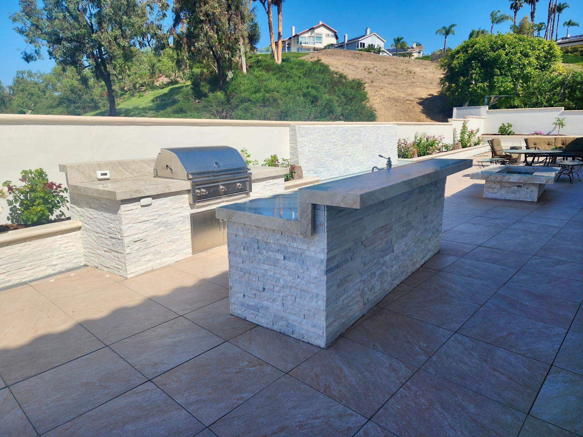 An outdoor kitchen with a stone-clad island, stainless steel grill, and tiled patio against a hillside landscape.