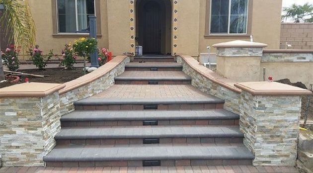 Wide stone steps with built-in lights lead to a home entrance, framed by tan stucco walls and stone-clad pillars.