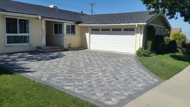 A light yellow, single-story house with a white garage door and a patterned grey paver driveway in a suburban setting.