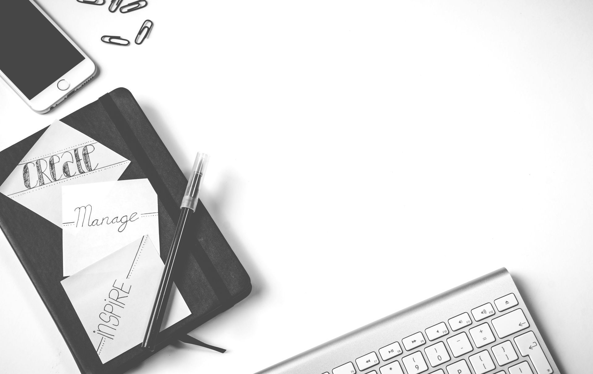 A black and white photo of a desk with a notebook , pen , cell phone and keyboard.