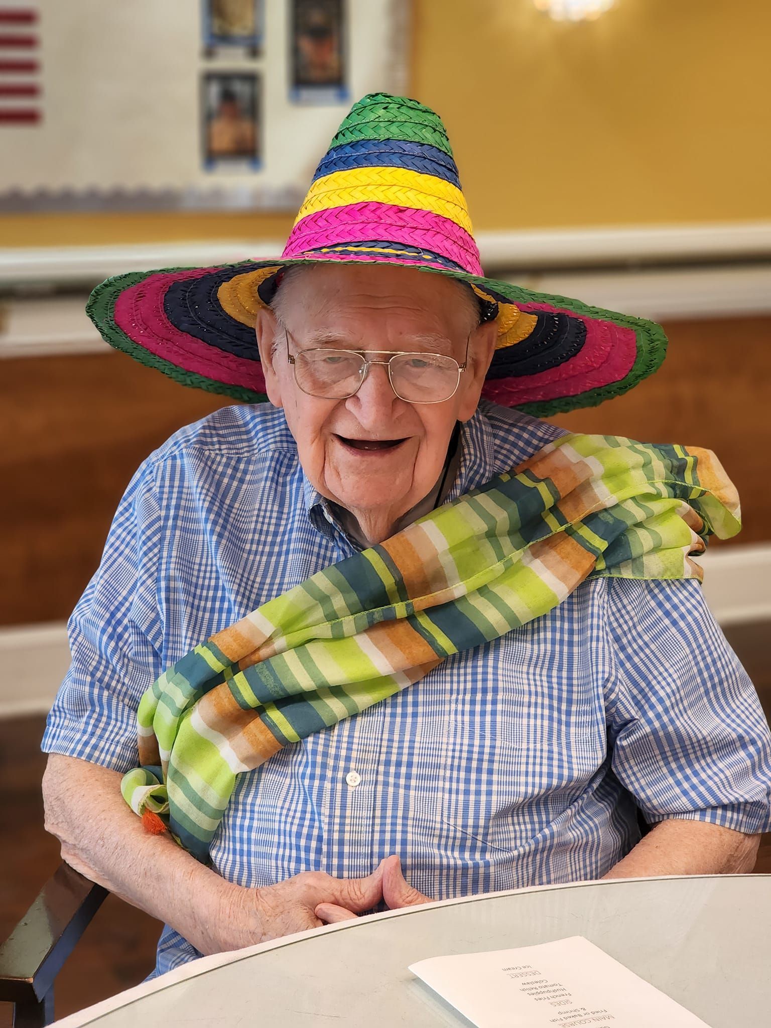 An elderly man wearing a sombrero and scarf is sitting at a table.