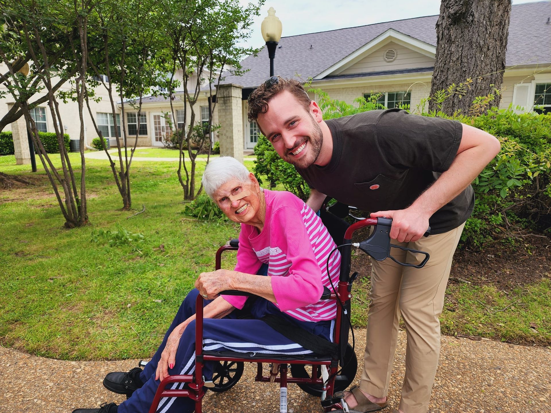 Two elderly women in wheelchairs are sitting on a patio while a man pushes a walker.
