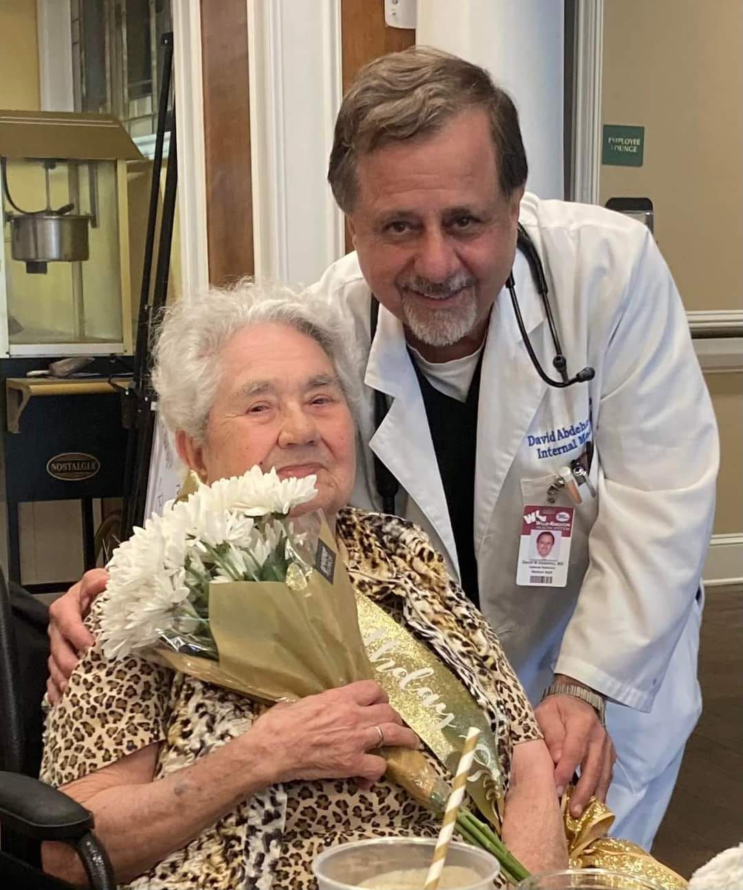 A man in a lab coat is standing next to an elderly woman holding a bouquet of flowers