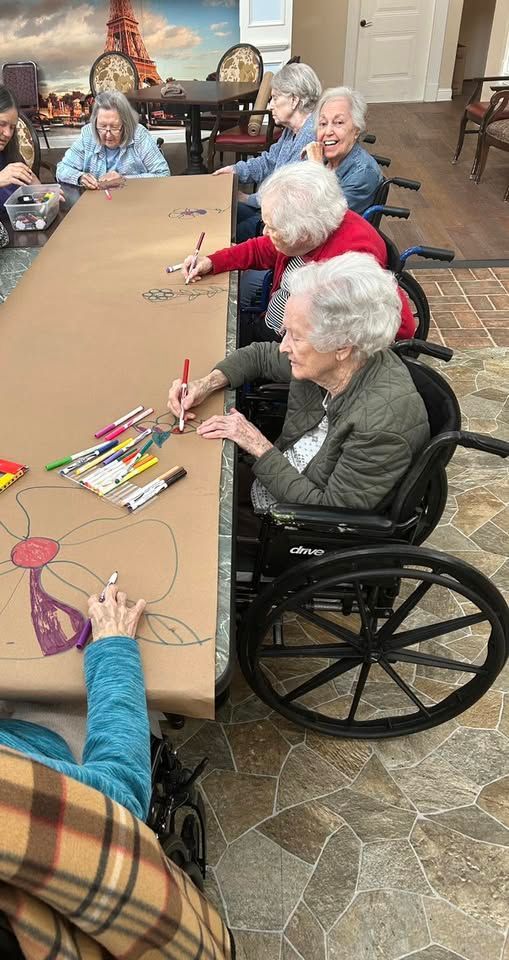 A group of elderly women in wheelchairs are sitting at a table drawing with markers.