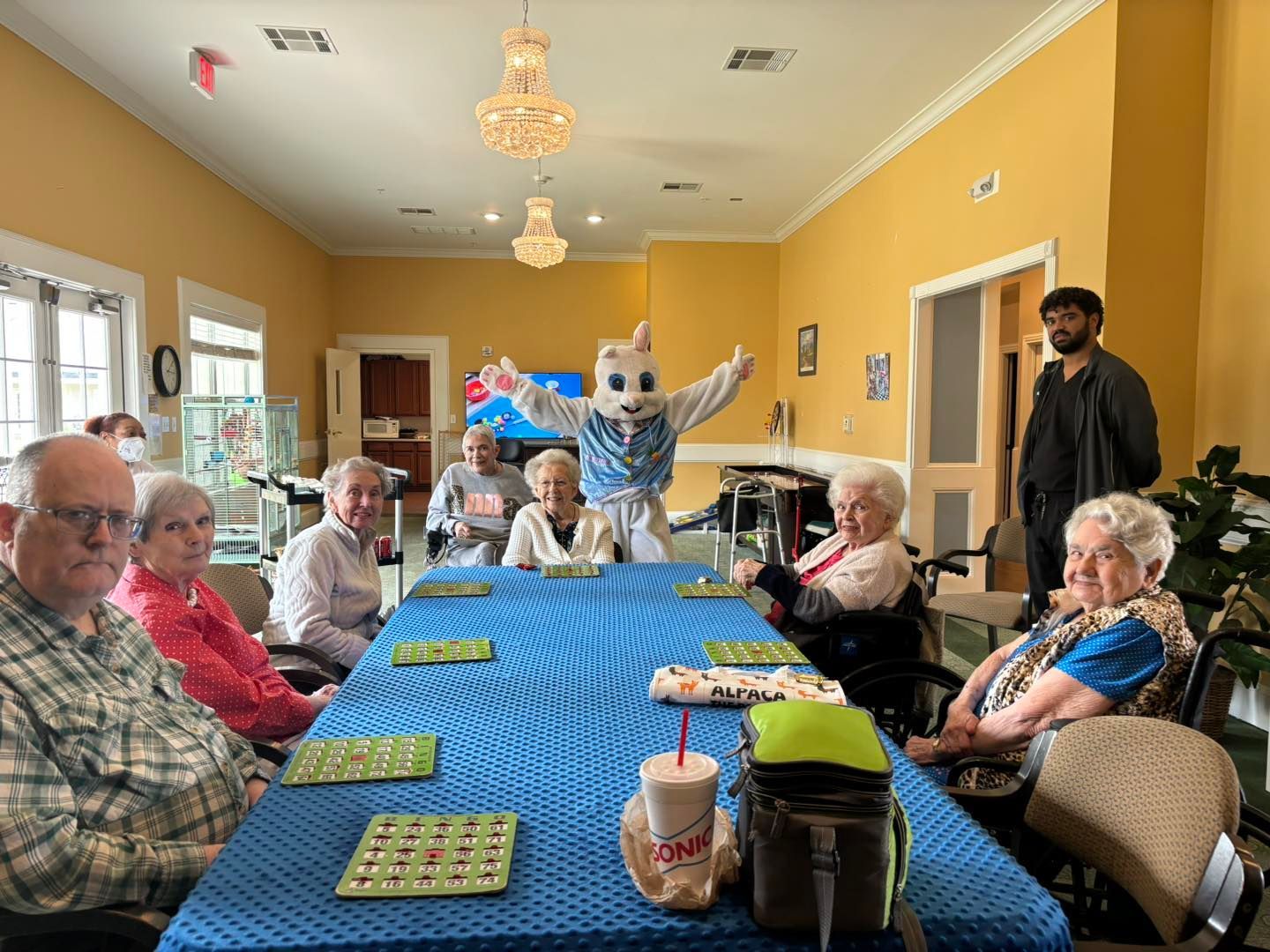 A group of elderly people are sitting around a table with a mascot in the background.