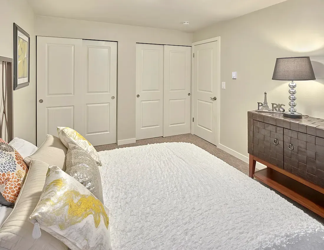A bright bedroom with a white shaggy rug, a bed with patterned pillows, white closet doors, and a decorative console.