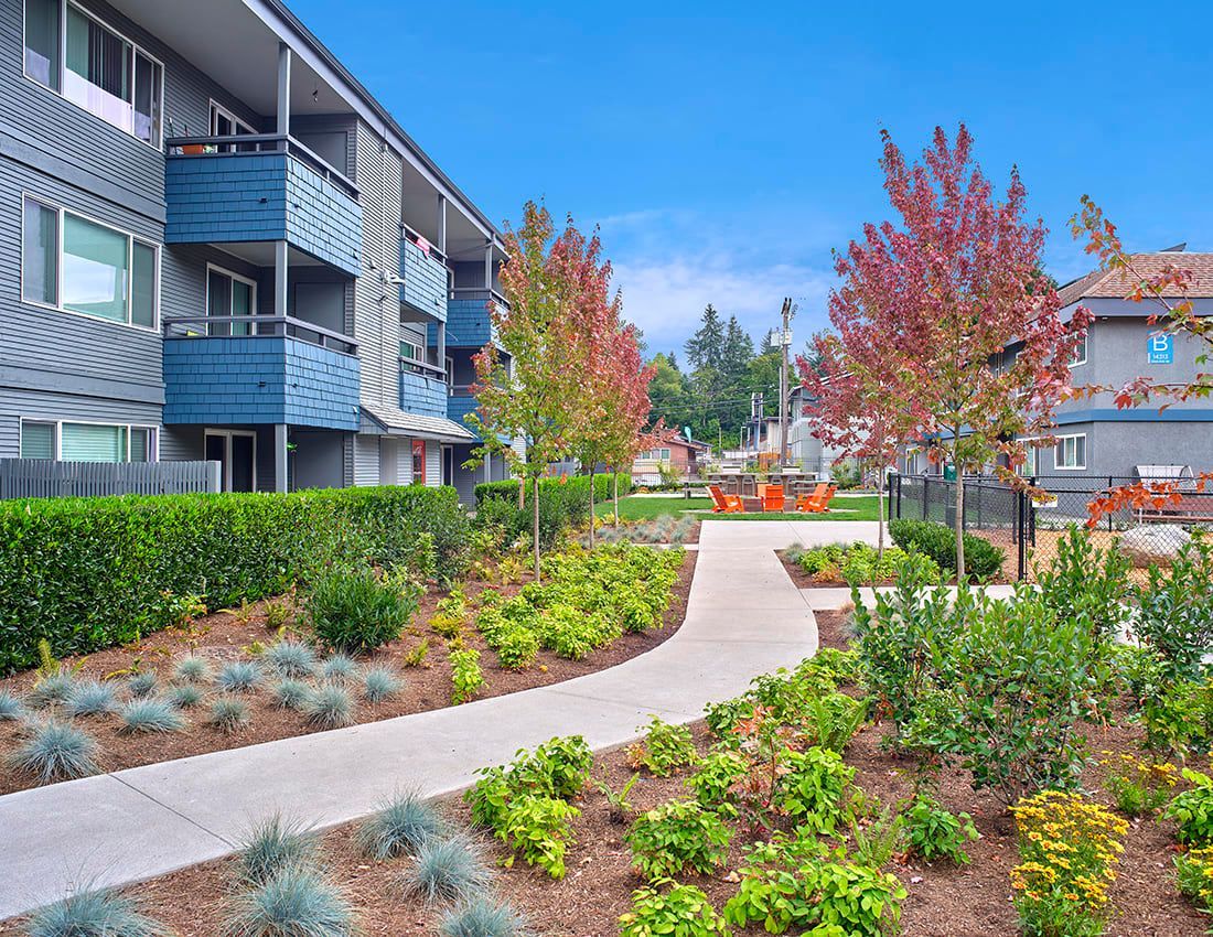 A paved path curves through a garden with shrubs and trees alongside a multi-story apartment building under a blue sky.