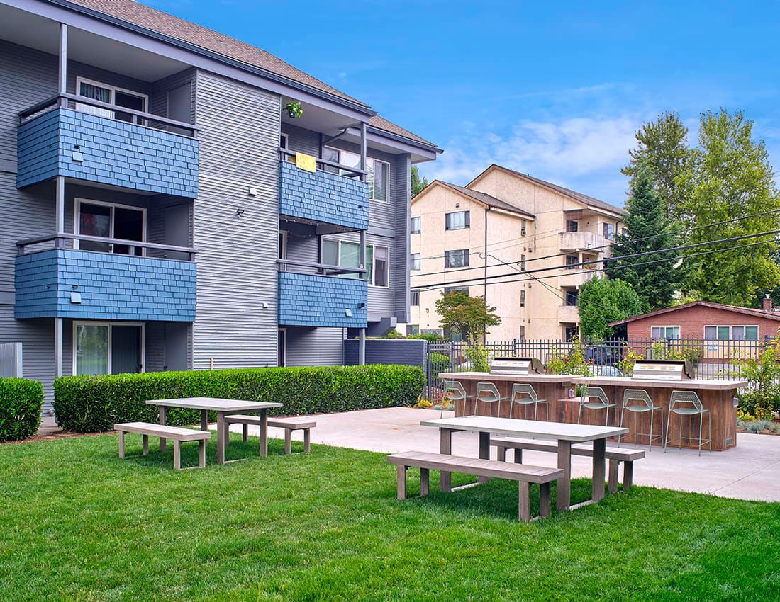 An outdoor apartment courtyard with picnic tables on a lawn and a paved area with bar-style seating and a grill.