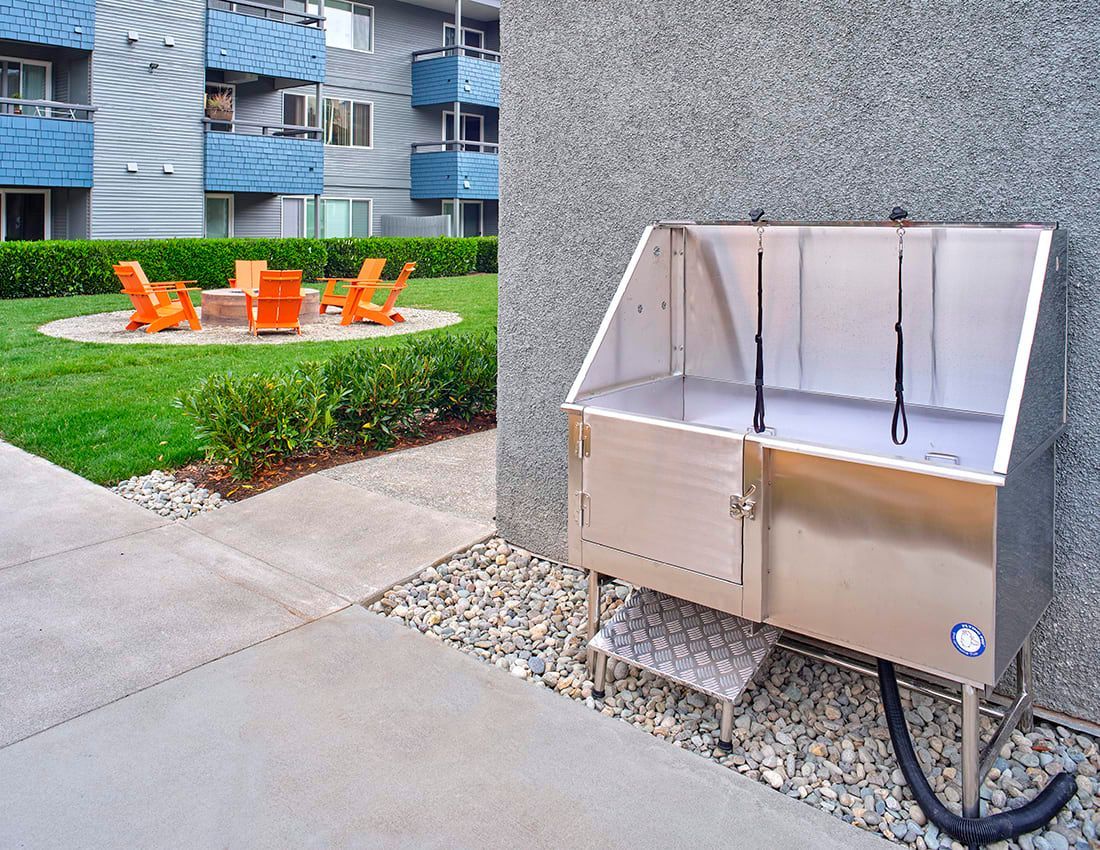 A stainless-steel outdoor pet washing station sits next to an apartment courtyard with orange chairs and a fire pit.