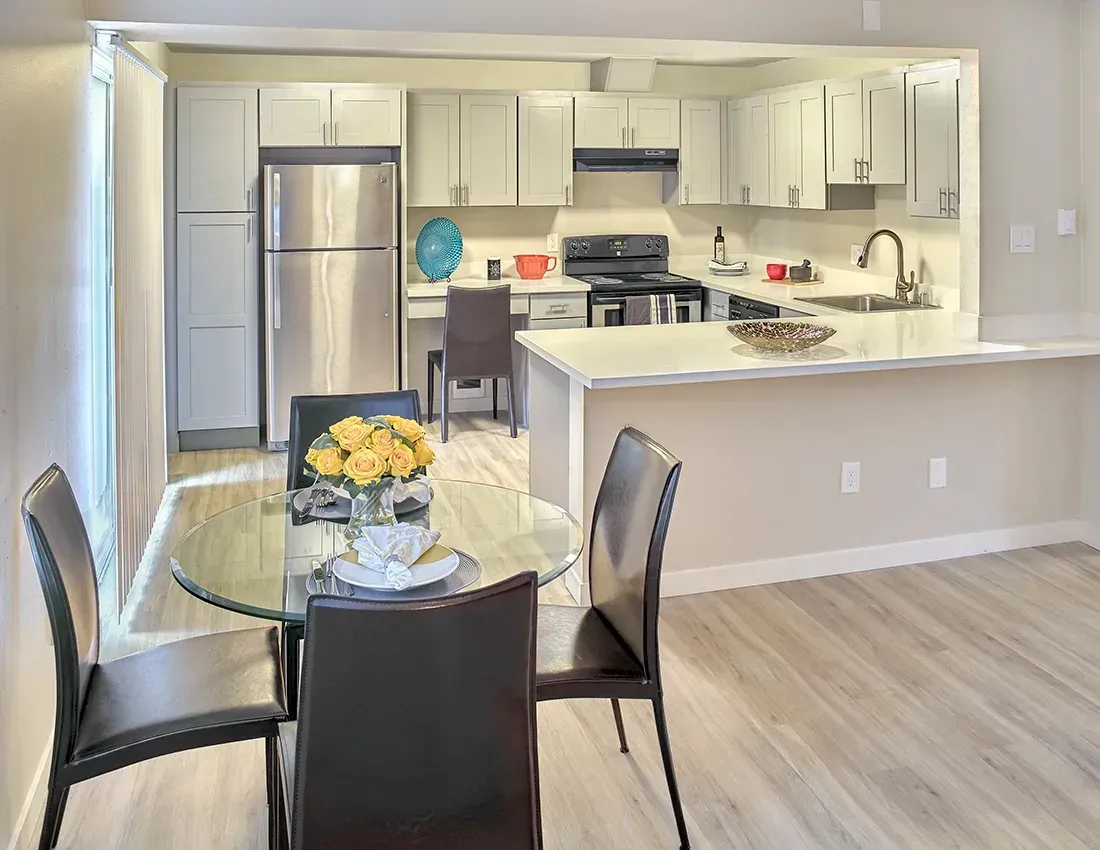A bright, open-plan kitchen and dining area with light wood floors, a glass table, and stainless steel appliances.