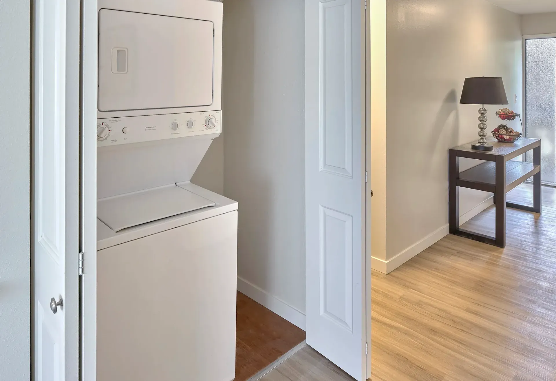 A stackable washer and dryer set inside a closet, next to a hallway with a side table, lamp, and light wood flooring.
