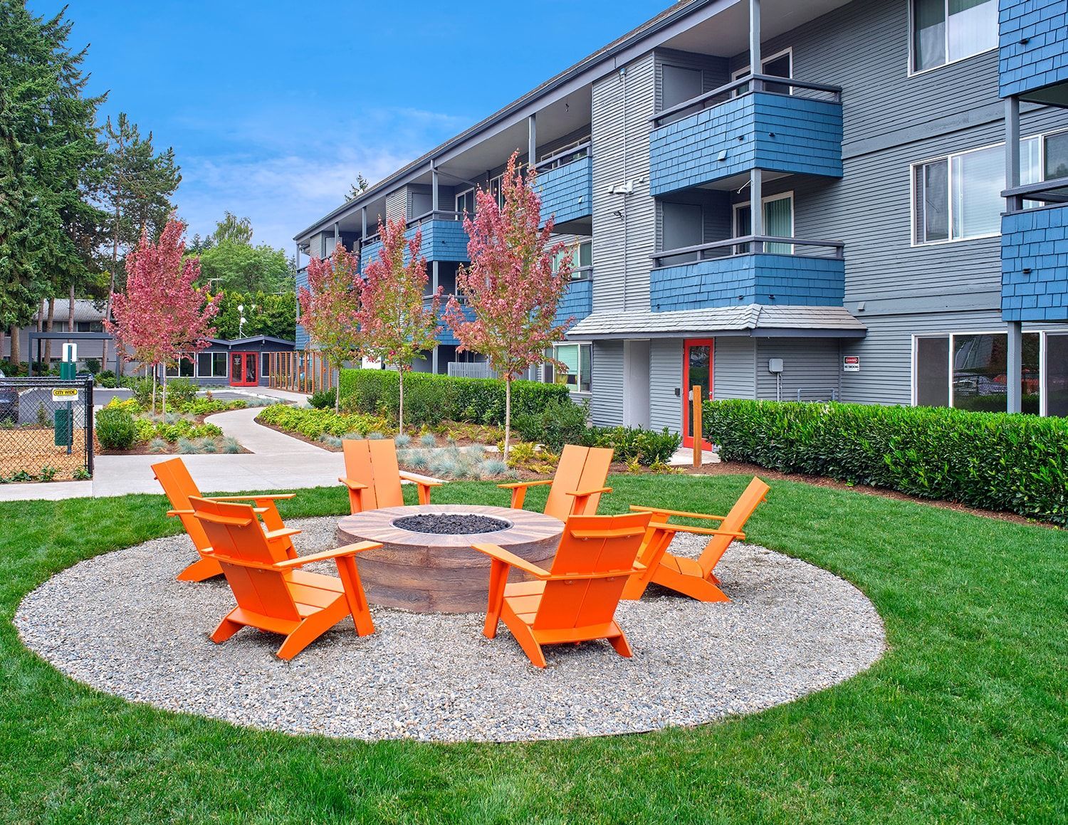Orange chairs surround a circular stone fire pit on a gravel patio outside a multi-story gray apartment building.