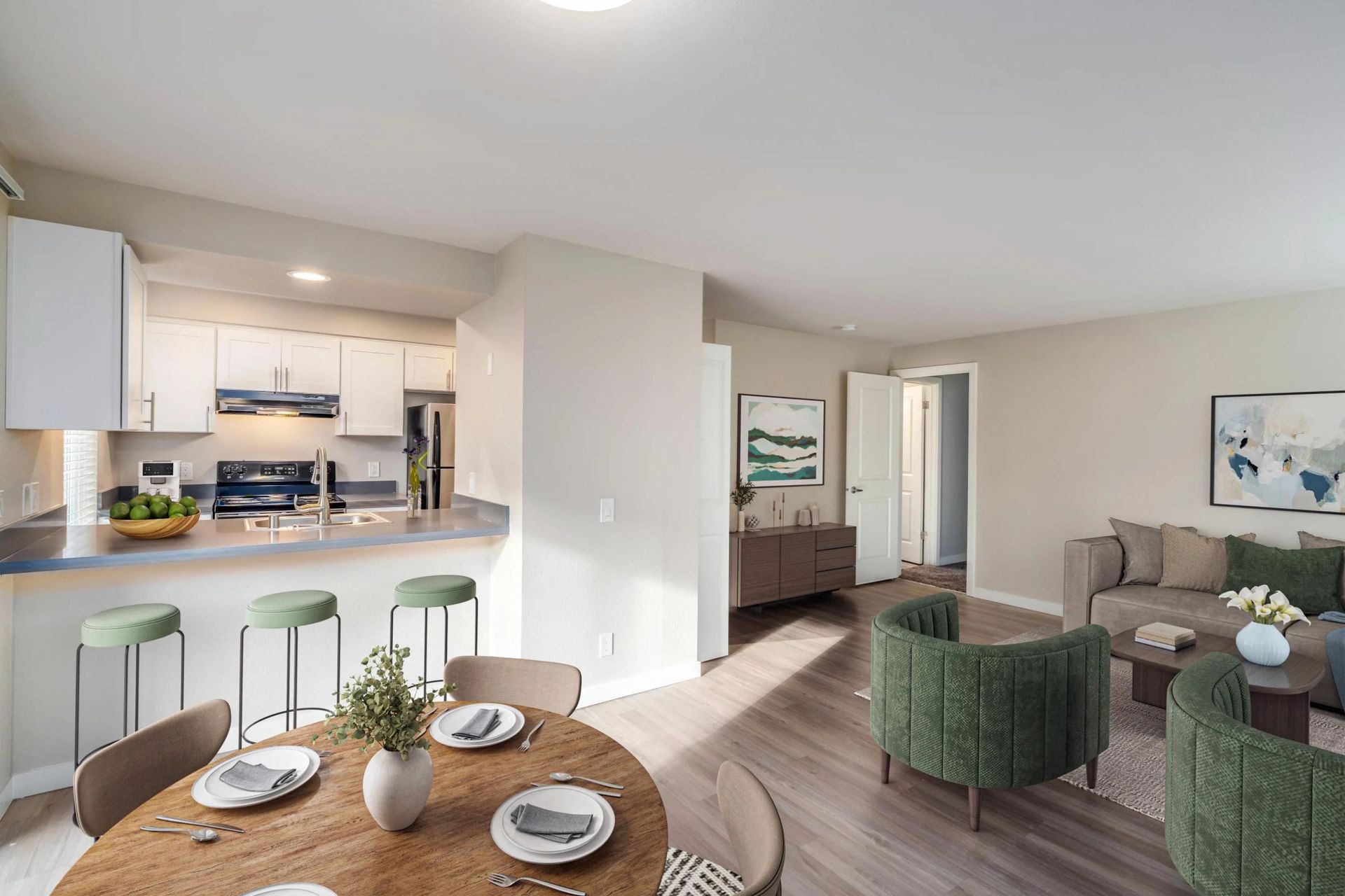 An open-concept apartment with a kitchen featuring white cabinets, a dining area, and a living room with green armchairs.