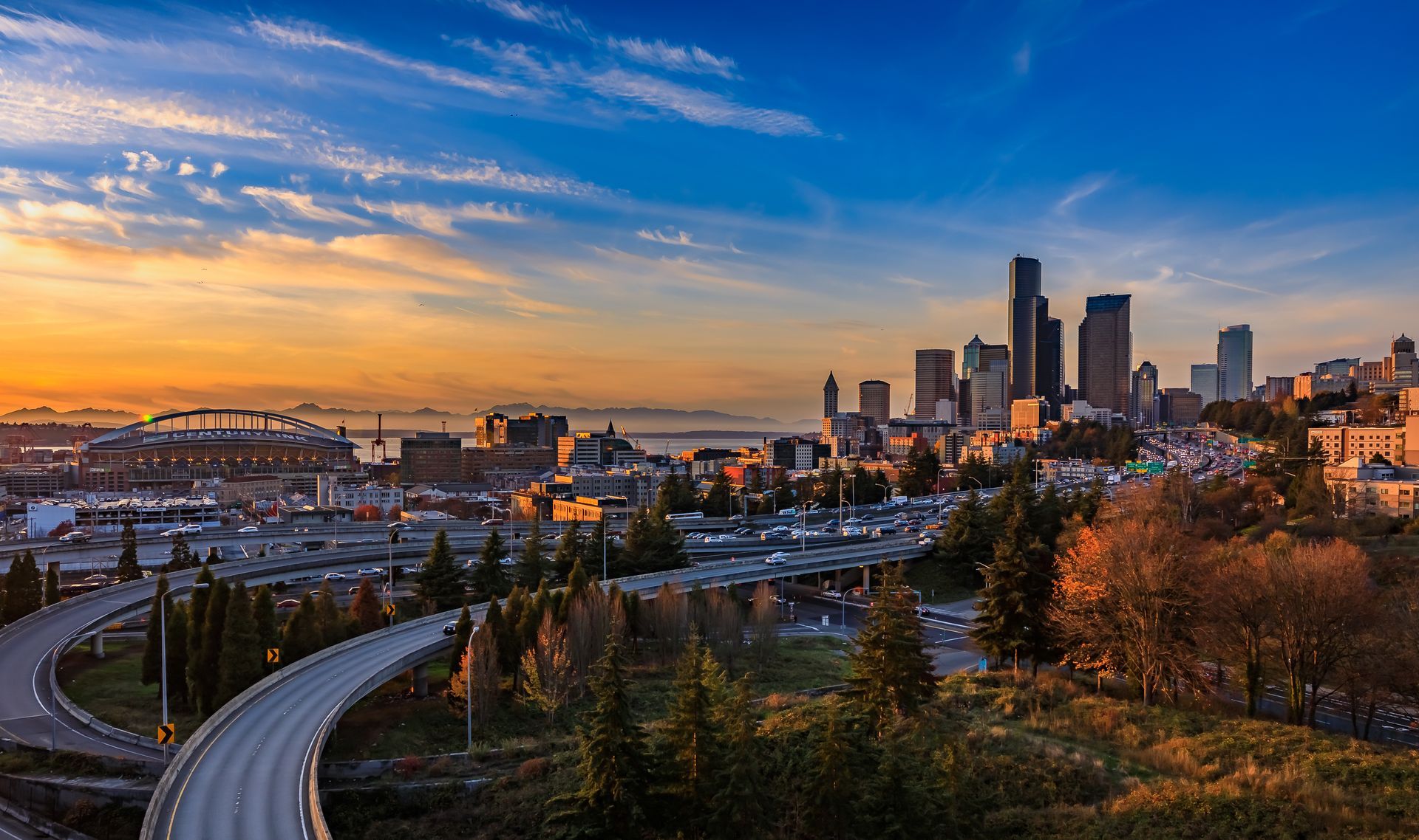 Seattle cityscape featuring the skyline, highway ramps, and stadium under a vibrant orange and blue sunset sky.
