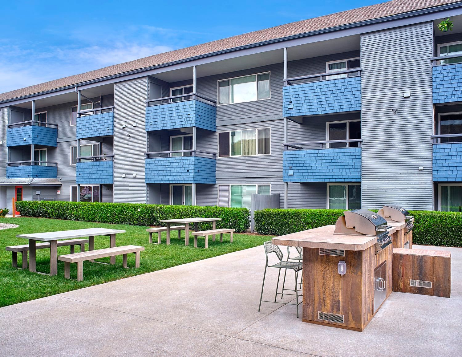 A three-story apartment building with a patio featuring a grill station, seating, and a picnic area on a grassy lawn.