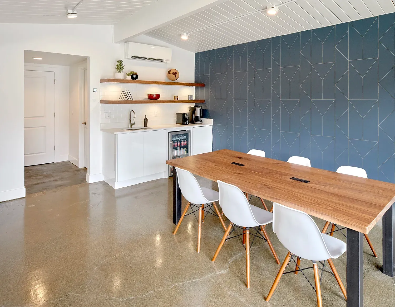 A modern meeting room with a wooden table, white chairs, kitchenette, and a dark blue geometric accent wall.