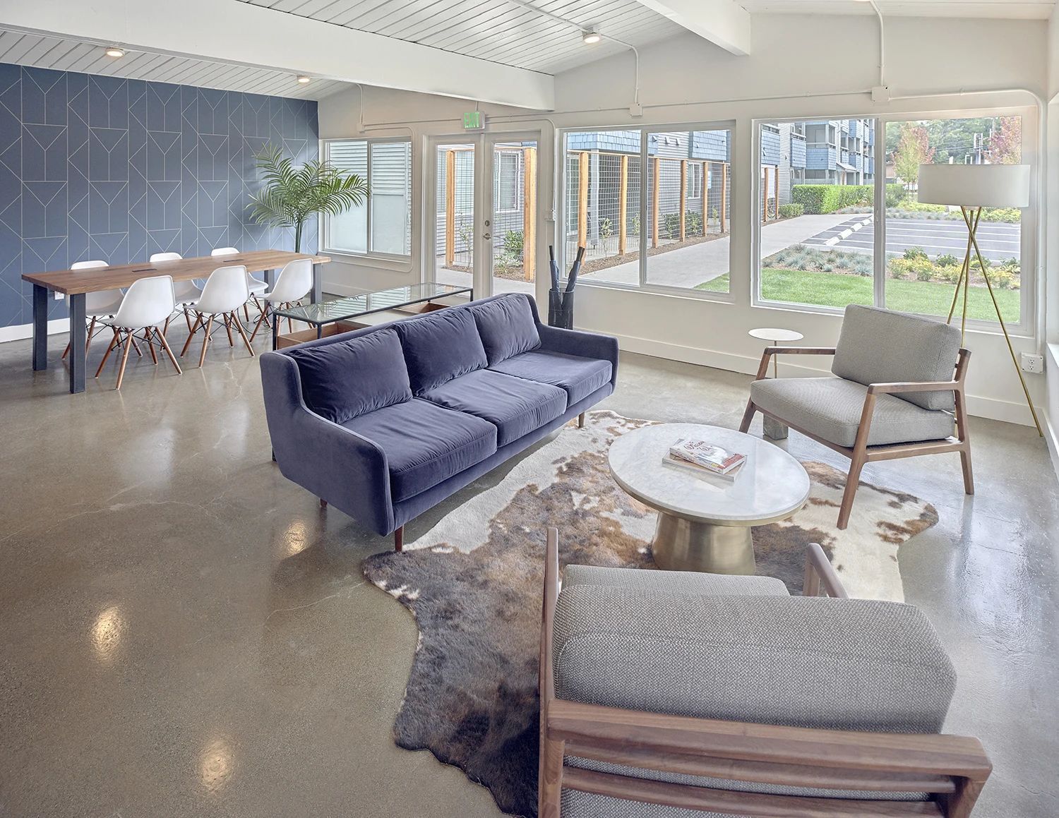 A modern lounge with a blue sofa, armchairs on a hide rug, coffee table, and a dining area by large windows.