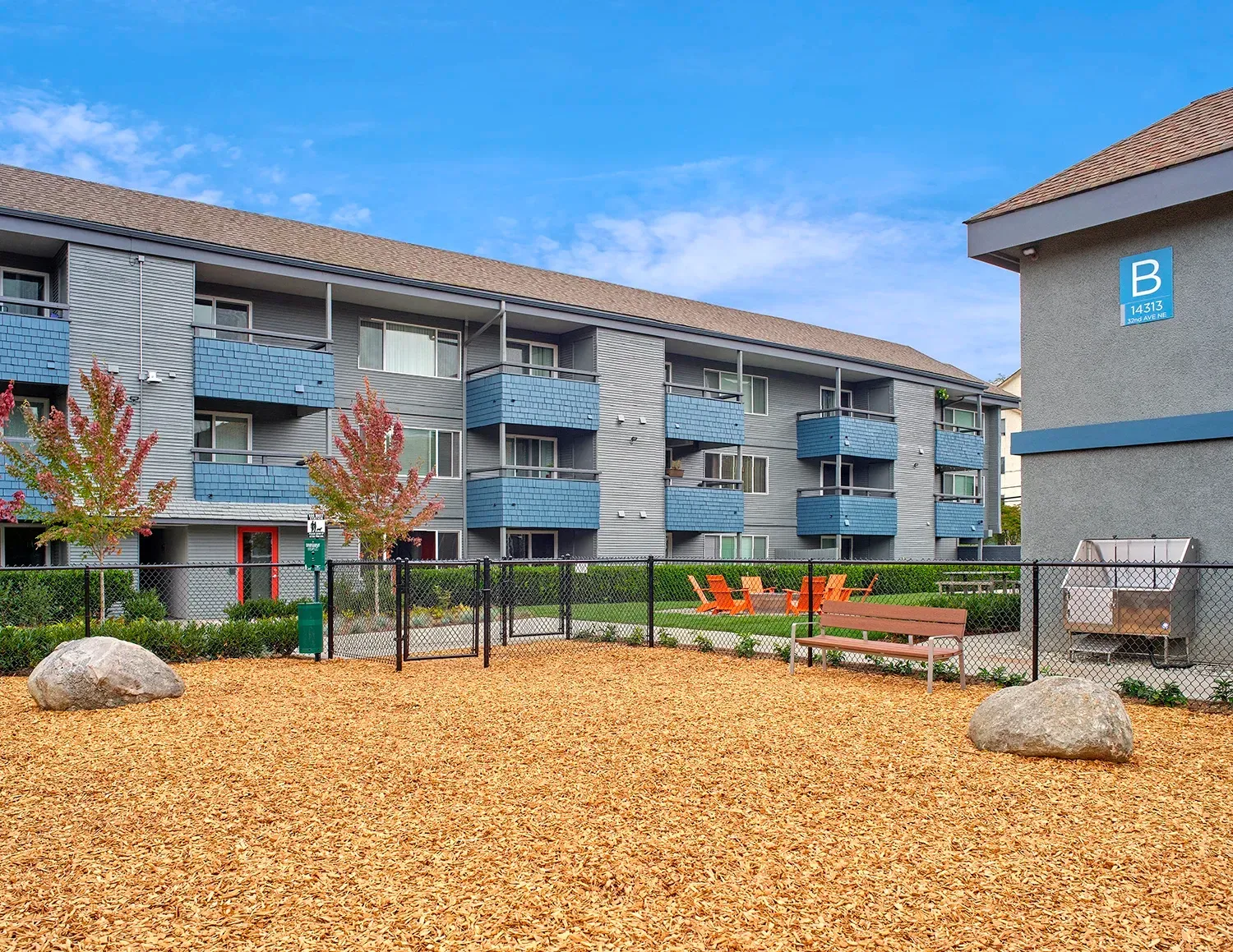 A dog park with wood chips, two large rocks, a bench, and a modern apartment building with blue balconies in the back.