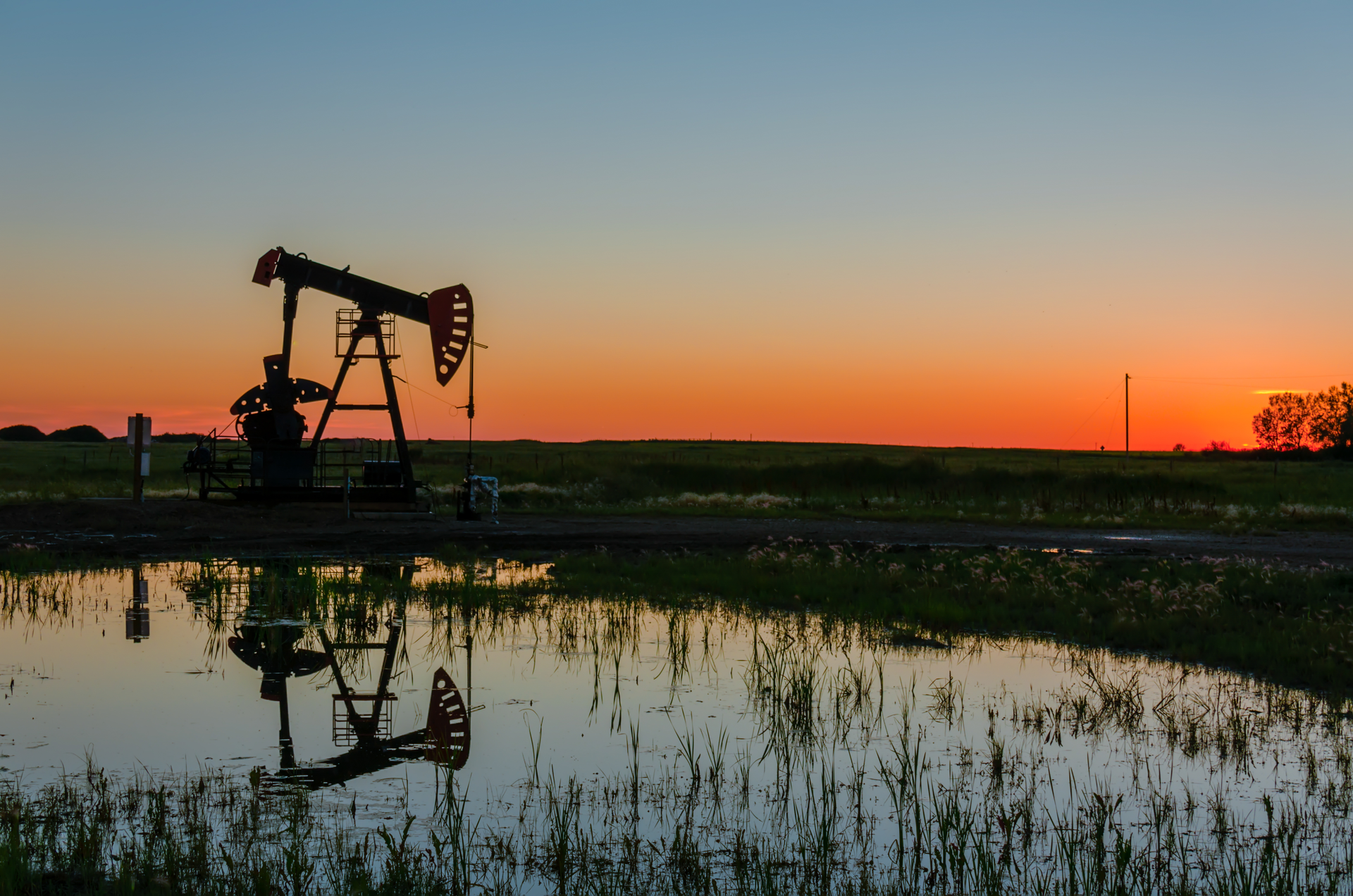 Oil pump jack at sunset reflecting on water in a rural field landscape, showing energy production scene.