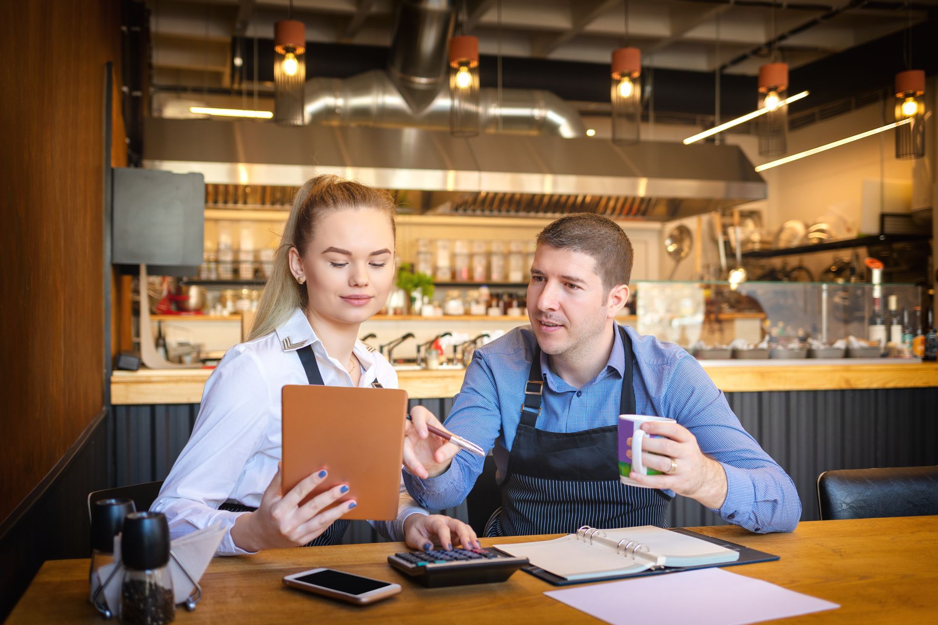 Woman and man in aprons review a tablet and notebook at a restaurant table.