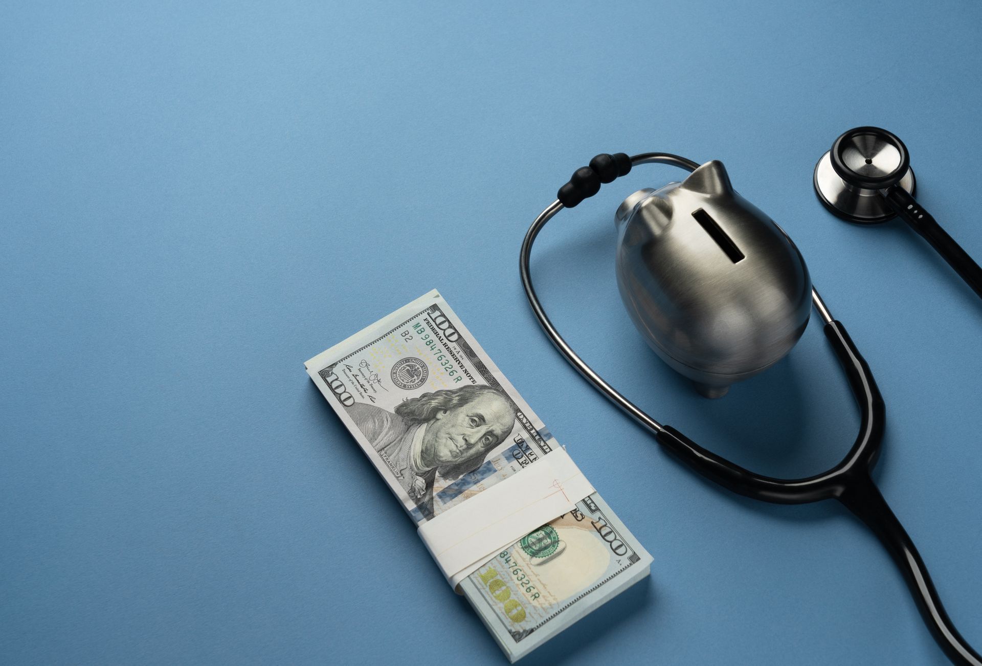 Stack of $100 bills, silver piggy bank, and stethoscope on blue background, suggesting healthcare finance.
