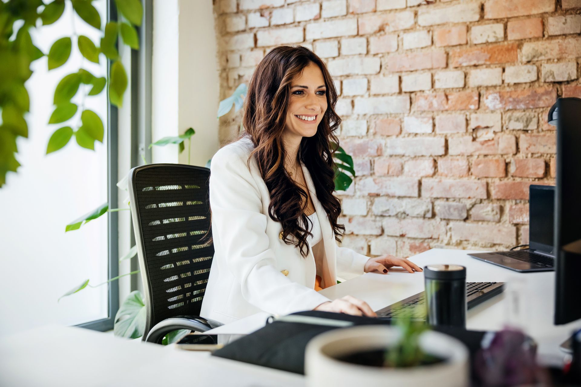 Woman in white blazer smiling, working at a desk with a computer, brick wall background.
