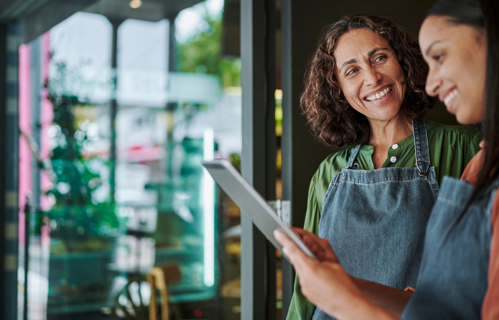 Two people looking at a tablet inside a business, one smiling, wearing an apron.