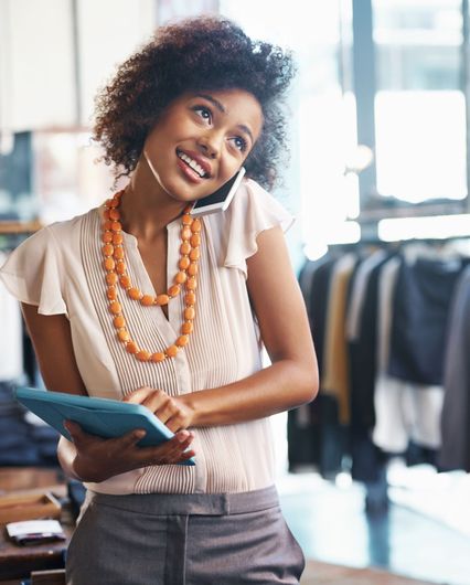 Woman in shop, smiling, using tablet while talking on phone, orange necklace.